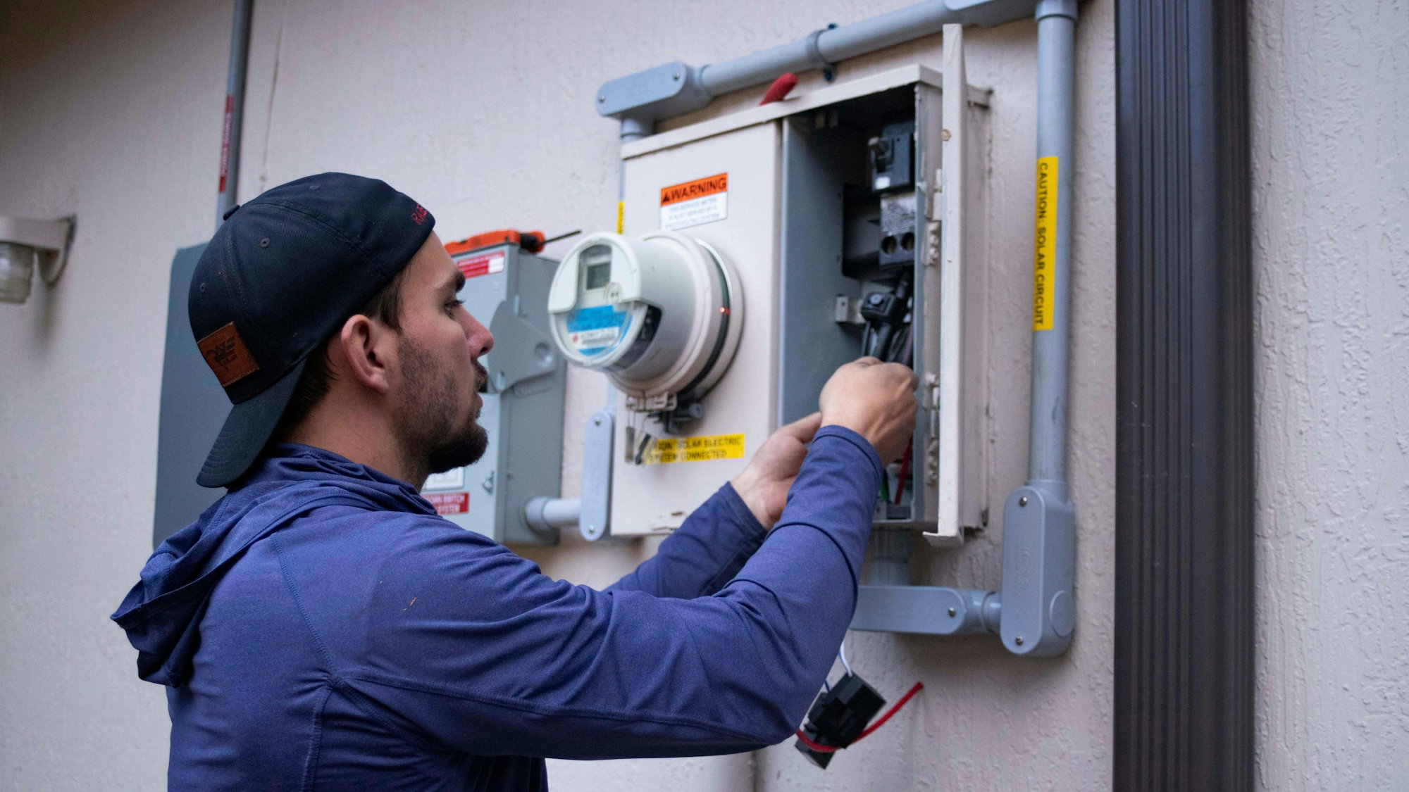a-person-in-a-blue-hoodie-and-baseball-cap-working-on-an-electrical-meter.jpg A person in a blue hoodie and baseball cap working on an electrical meter